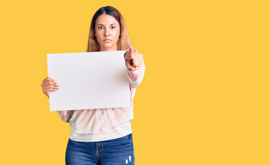 Fototapeta na wymiar Beautiful young woman holding blank empty banner pointing with finger to the camera and to you, confident gesture looking serious