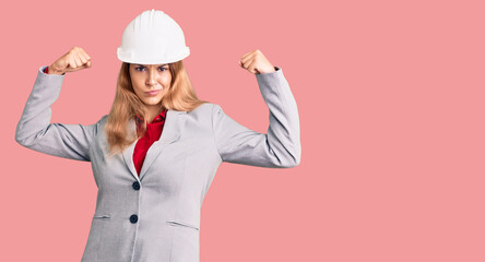 Beautiful young woman wearing architect hardhat showing arms muscles smiling proud. fitness concept.