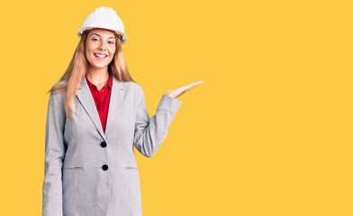 Beautiful young woman wearing architect hardhat smiling cheerful presenting and pointing with palm of hand looking at the camera.