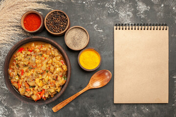 top view roasted eggplant salad in bowl different spices in small bowls notebook wooden spoon on dark background