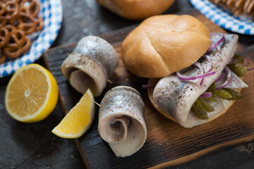 Fototapeta premium German fish sandwich with herring filet, pickles, red onion and lemon on a rustic wooden cutting board, closeup