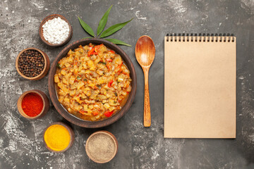 top view roasted eggplant salad in bowl different spices in bowls wooden spoon leaf notepad on dark background