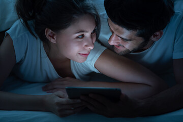 Happy young couple looking a tablet together and laughing while lying on the bed at night