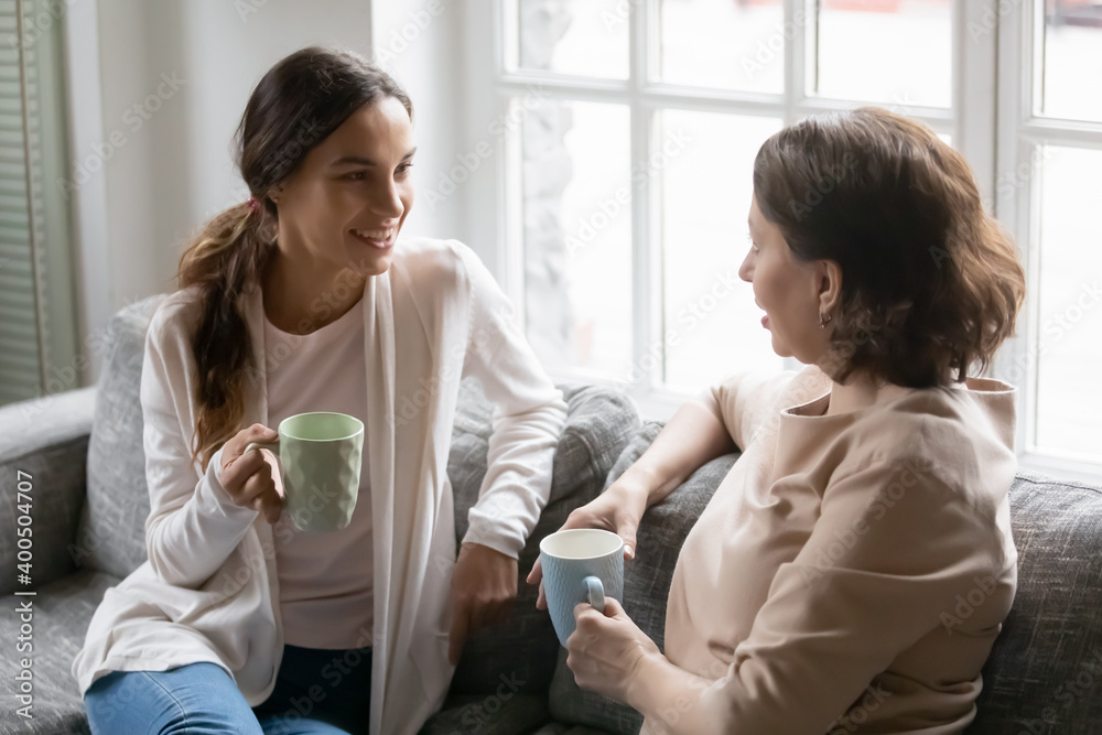 Wall mural Close up happy grownup daughter and mature mother chatting, spending weekend together, sitting on cozy couch, holding cups of hot drinks, grandmother and granddaughter enjoying pleasant conversation