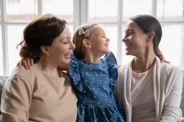 Obraz premium Close up overjoyed three generations of women having fun together, cuddling and laughing, cute little girl hugging young mother and mature grandmother, family enjoying leisure time at home