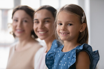 Head shot portrait happy three generations of women standing in row, aging process concept, focus on smiling little girl looking at camera, young mother and mature grandmother on background