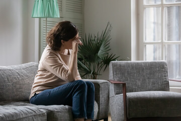 Depressed pensive middle aged woman lost in thoughts, sitting on couch at home alone, looking in distance, upset mature female thinking about problems, touching forehead, suffering from headache