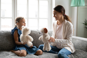 Close up happy young mother and little daughter playing with fluffy toys, sitting on cozy couch at...