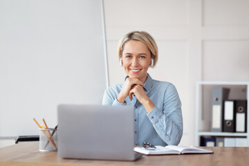 Portrait of young teacher sitting near laptop, looking at camera and smiling, giving online lesson,...