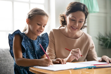 Obraz premium Close up happy middle aged grandmother and little granddaughter drawing pencils, sitting at table, smiling mature woman babysitter teacher and cute girl child engaged in creative activity