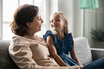 Close up happy mature woman with cute little girl talking, sharing news, smiling grandmother and granddaughter chatting, sitting on cozy couch at home, spending leisure time, weekend together