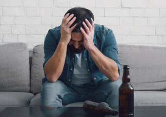 Sad alcoholic holds his head with hands, empty bottles on table