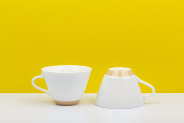 Two white glossy ceramic cups on white table against bright yellow background with a space for text
