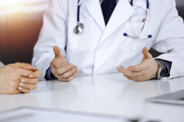 Unknown male doctor and patient woman discussing something while sittingin a darkened clinic, glare of light on the background. Close-up of hands