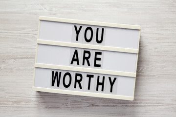 'You are worthy' on a lightbox on a white wooden background, top view. Flat lay, overhead, from above.