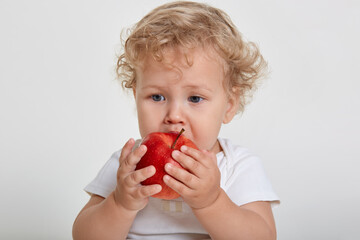Baby with apple posing isolated over white background, blond tot biting red fruit and looking away, vitamins for male kid, wavy haired child having snack.