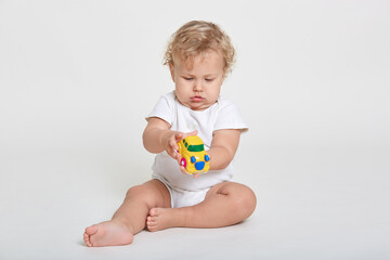 Fototapeta na wymiar Curious kid playing with toy while sitting on floor, wearing bodysuit, posing barefoot, having wavy blond hair, cute toddler having fun indoor, holds toy with both hands.