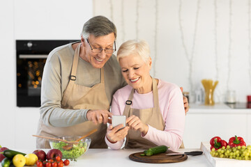Happy Senior Couple Using Smartphone Preparing Dinner Standing In Kitchen