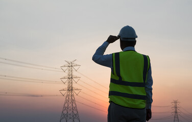 An Asian male engineer works in front of a high-voltage tower. In the sunset the sky is beautiful