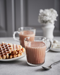 Hot cocoa drink on a kitchen table in two glass mugs, served with freshly baked waffles and some sugar. Porcelain flower bouquet as a table decoration on the background. Blurred kitchen panels behind