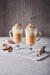 Two pumpkin spice lattes in tall latte glasses, topped with whipped cream and chocolate dust, on light grey table. Near them: ground spices in glass bowls, whole nutmegs, small grater, cinnamon sticks
