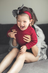 Obraz na płótnie Canvas A toddler wearing a red top and a hair band with a hair bow laughs and plays on top on her mother’s legs as she tickles her while sitting on a sofa in a house in Edinburgh, Scotland, United Kingdom