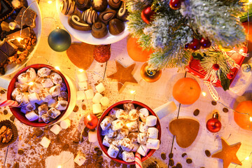 sweet food top view background for merry christmas or new year holiday decoration with night illumination - chocolate candies, tangerines, cookies, marshmallow and cocoa latte on white wood