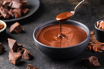 Hot melted chocolate dripping from a spoon into a bowl of chocolate on a dark textured background....