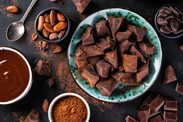 Chunks of dark chocolate on a plate, melted chocolate in a bowl and cocoa beans on a dark textured background. Chocolate confectionery background.