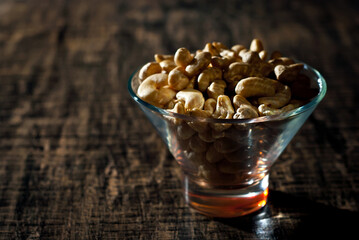 Cashews in a clear glass bowl on an old shabby board. Nuts on a brown wooden table. Contrasting dramatic light as an artistic effect.
