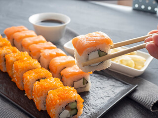 Philadelphia and California rolls on a textured dark plate. One roll is held in Granny sticks above the others. Next to soy sauce, wasabi and ginger. Side view. Japanese cuisine