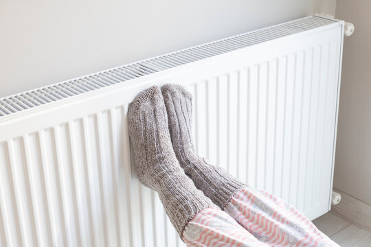 Woman is warm knitted woolen socks near a home heater in cold winter time. Using heater at home in winter.