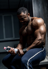 Fototapeta na wymiar African athlete in the locker room with bottle of water. Strong man with perfect body and strong abs. Rest after training.