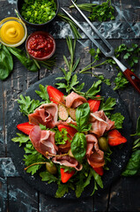 Salad of arugula, prosciutto, strawberries and capers on a black stone plate. Italian cuisine.