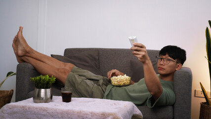 Portrait of asian man in casual watching interesting movie on TV and eating popcorn while sitting on the couch in living room at home, people and entertainment concept.