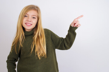 Caucasian kid girl wearing green knitted sweater against white wall looking at camera indicating finger empty space sales