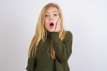 Shocked Caucasian kid girl wearing green knitted sweater against white wall looks with great surprisment being very stunned, astonished with unexpected news, Facial expressions concept.
