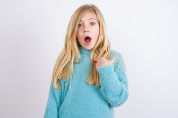 Cute Caucasian kid girl wearing blue knitted sweater against white wall being in stupor shocked,...