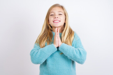 Cute Caucasian kid girl wearing blue knitted sweater against white wall keeps palms together, has...