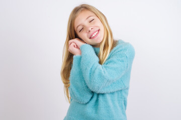 Dreamy Cute Caucasian kid girl wearing blue knitted sweater against white wall with pleasant expression, closes eyes, keeps hands crossed near face, thinks about something pleasant