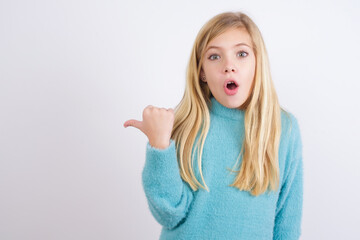 Shocked Cute Caucasian kid girl wearing blue knitted sweater against white wall points with thumb away, indicates something. Check this out. Advertisement concept.