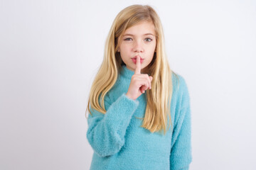 Cute Caucasian kid girl wearing blue knitted sweater against white wall makes hush gesture, asks be quiet. Don't tell my secret or not speak too loud, please!