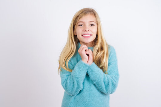 Dreamy charming Cute Caucasian kid girl wearing blue knitted sweater against white wall with pleasant expression, keeps hands crossed near face, excited about something pleasant.