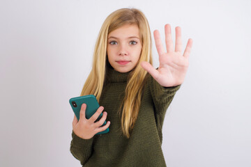 Caucasian kid girl wearing green knitted sweater against white wall using and texting with...