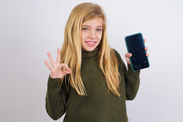 Caucasian kid girl wearing green knitted sweater against white wall  holding in hands cell showing...