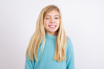 Cute Caucasian kid girl wearing blue knitted sweater against white wall blinking eyes with pleasure having happy expression. Facial expressions and people emotions concept.