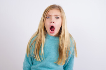 Expressive facial expressions. Shocked stupefied Cute Caucasian kid girl wearing blue knitted sweater against white wall, keeps jaw dropped feels stunned from what he sees aside.
