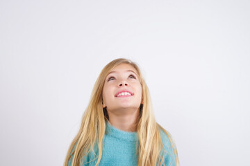 Cute Caucasian kid girl wearing blue knitted sweater against white wall looking up as he sees something strange.