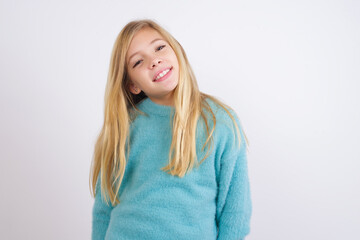 Cute Caucasian kid girl wearing blue knitted sweater against white wall with broad smile, shows white teeth, feeling confident rejoices having day off.