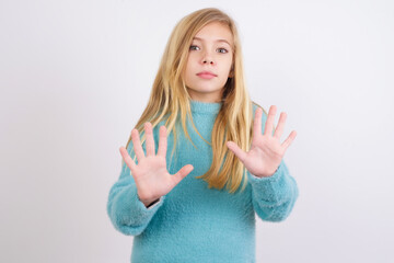 Cute Caucasian kid girl wearing blue knitted sweater against white wall doing stop sing with palm of the hand. Warning expression with negative and serious gesture on the face.